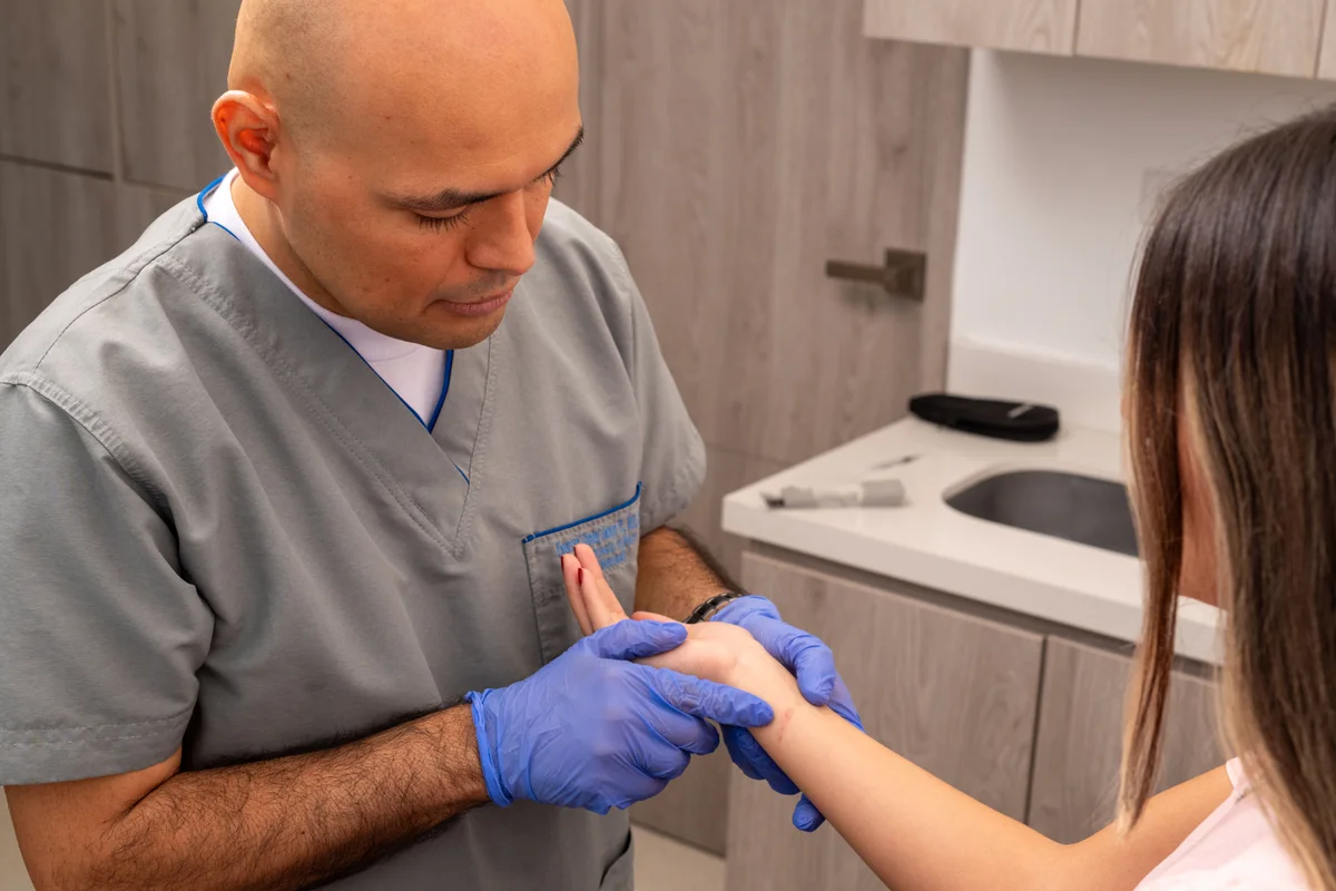 Dr. Daniel Amaya examinando con guantes clínicos lesión eccematosa en la muñeca de una paciente