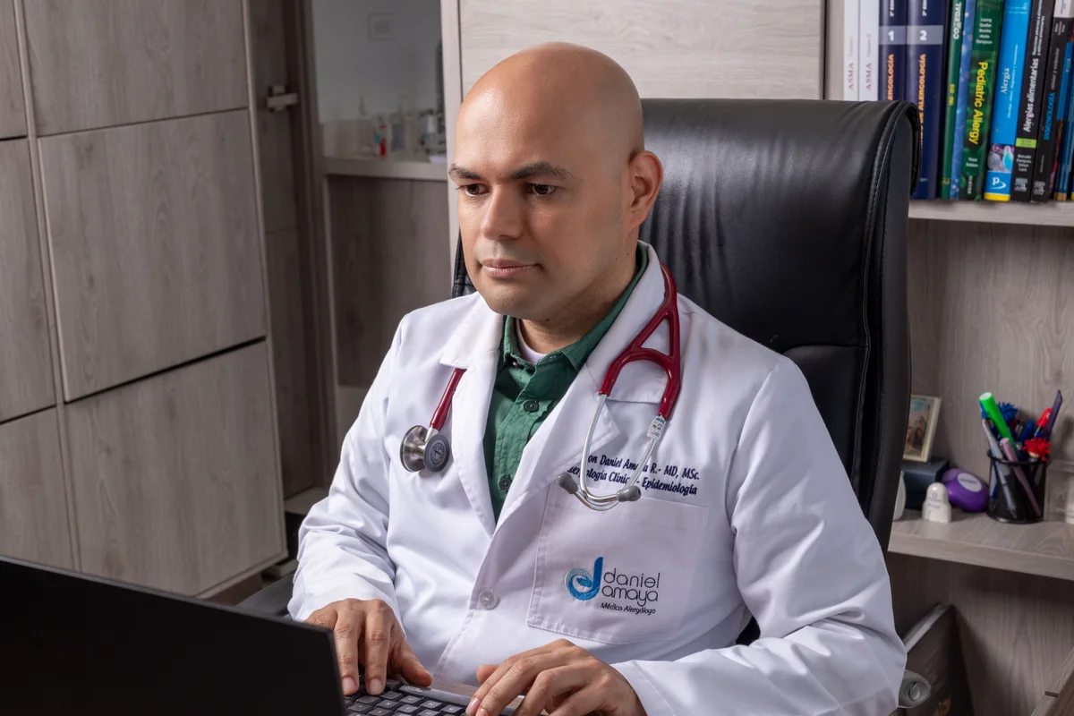 Dr. Daniel Amaya trabajando en su computador portátil en el consultorio, rodeado de literatura médica de alergología